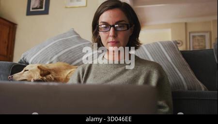 Eine Tippfrau mit Brille, gedämpftem grünen Pullover auf dem Sofa zu Hause, mit Laptop und schlafendem Hund Stockfoto