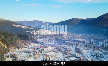 Winter Mountain Village Luftlandschaft. Blick aus der Vogelperspektive auf ein malerisches Bergdorf, umgeben von Kiefernwäldern und sanften Hügeln Stockfoto