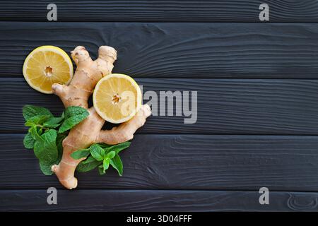 Ingwer, Lemon und Mint auf schwarzem hölzernem Hintergrund. Frische Ingwerwurzel, Zitronenscheiben und Minzblätter auf einer strukturierten schwarzen Holzoberfläche angeordnet. Oben Stockfoto