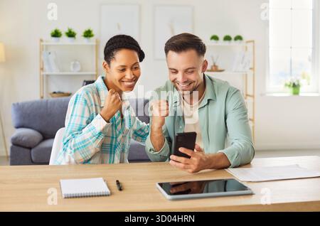Junges glückliches, aufgeregtes Paar, das zu Hause am Schreibtisch sitzt und auf das Mobiltelefon blickt und eine Ja-Geste macht Stockfoto