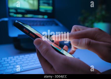 Nahaufnahme von Personen, die nachts mit Social-Media-Feed auf dem Smartphone-Bildschirm mit Laptop im Hintergrund aufzeichnen. Digitale sucht und moderne Online-Gewohnheiten. Stockfoto