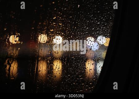 Regentropfen Rollen auf einem Autofenster mit verschwommenen, bokeh-Lichtern im Hintergrund bei Nacht Stockfoto