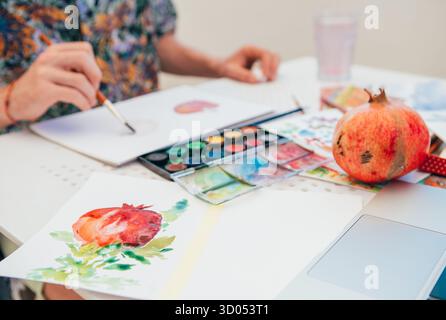 Während der kreativen Hobbysession konzentriert sich der junge Mann darauf, Granatapfelfrüchte mit Pinsel und Palette in ihrem Skizzenblock zu malen. Nahaufnahme hebt das hervor Stockfoto
