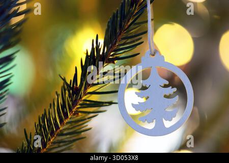 Weißes, weihnachtbaumförmiges hölzernes Ornament, das an einem Weihnachtsbaum hängt, mit bunten Bokeh-Lichtern im Hintergrund Stockfoto