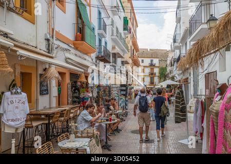 Menschen schlendern durch eine bezaubernde, enge Straße, die mit kleinen Geschäften und Außentreffen gesäumt ist, im Zentrum von Ibiza-Stadt, Spanien. Stockfoto