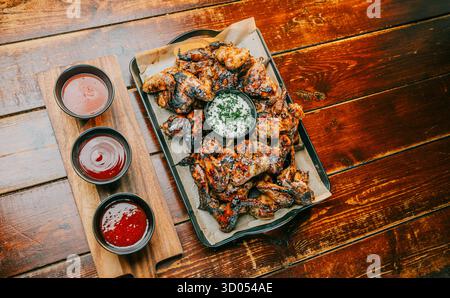 Auf einem Holztisch mit drei Gewürzbechern wird ein Tablett mit Hähnchenflügeln serviert. Die Würzmittel sind Ketchup, Senf und Genießer. Der Tisch besteht aus W Stockfoto