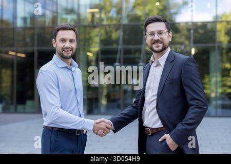 Zwei Geschäftsleute in Anzügen lächeln und schütteln vor einem modernen Bürogebäude die Hände, schließen eine Partnerschaftsvereinbarung ab und feiern eine erfolgreiche professionelle Zusammenarbeit und einen erfolgreichen Abschluss Stockfoto