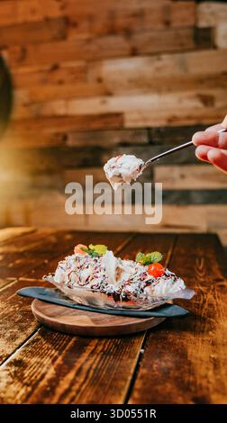 Eine Person isst ein Dessert mit einer Gabel. Das Dessert ist ein Bananenaufschnitt mit Eis und Belag. Das Konzept von Genuss und Genuss als Person Stockfoto