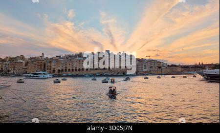 Warmer Sonnenuntergang über dem Hafen von Senglea Isla, historische Uferpromenade und vertäute Boote, weiche Wolken und Pastellstrahlen am mediterranen Himmel: Birgu, Malta - Octo Stockfoto