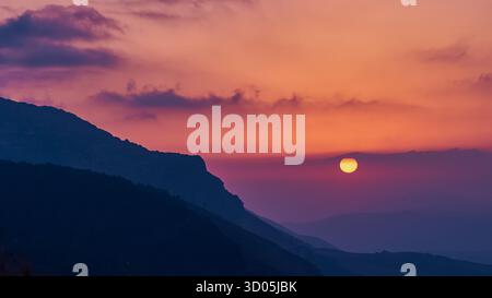 Warme Sonne untergeht hinter trüben Bergkämmen, rosa und violette Wolken am ruhigen Abendhimmel, ruhige stimmungsvolle Landschaft Stockfoto