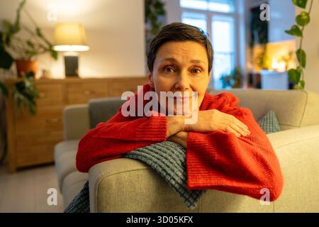 Lächelnde reife Frau, die Kopf auf Händen ruht. Glückliche, weibliche Kamera auf dem Sofa, anmutiger Ruhestand Stockfoto