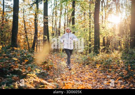 Frau mittleren Alters, die durch den Herbstwald joggt und einen aktiven Lebensstil inmitten farbenfroher Laub genießt. Morning Fitness Routine Outdoor Wellness, h Stockfoto
