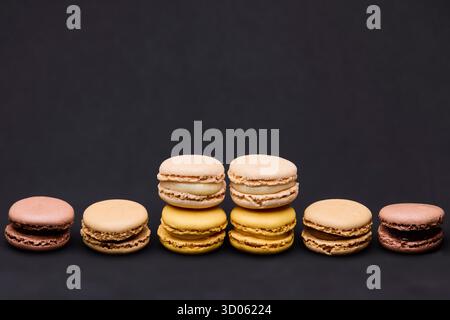 Eine Linie von fünf farbenfrohen Macarons auf einem schlichten dunklen Hintergrund. Die minimale Zusammensetzung betont Muster, Symmetrie und Textur für die Dessertwerbung Stockfoto
