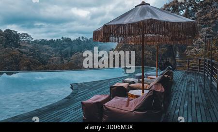 Ruhiger Infinity-Pool mit Liegestühlen und Sonnenschirm mit Blick auf den tropischen Regenwald an einem bewölkten Tag in Bali, Indonesien. Stockfoto