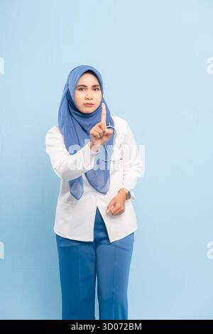 Porträt eines ernsthaften asiatischen muslimischen Arztes oder einer Krankenschwester, der mit den Fingern eine „Stop“- oder „No“-Geste zeigt, isoliert auf blauem Hintergrund. Stockfoto