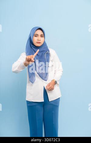 Porträt eines ernsthaften asiatischen muslimischen Arztes oder einer Krankenschwester, der mit den Fingern eine „Stop“- oder „No“-Geste zeigt, isoliert auf blauem Hintergrund. Stockfoto