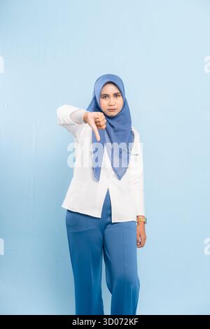 Porträt eines ernsthaften jungen asiatischen muslimischen Arztes oder Krankenpflegers, der mit den Daumen nach unten zeigt, was auf Missbilligung oder negative Prognose hindeutet, isoliert auf einem blauen Hintergrund Stockfoto