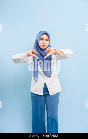 Porträt eines ernsthaften jungen asiatischen muslimischen Arztes oder Krankenpflegers, der mit den Daumen nach unten zeigt, was auf Missbilligung oder negative Prognose hindeutet, isoliert auf einem blauen Hintergrund Stockfoto