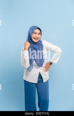 Porträt eines selbstbewussten asiatischen muslimischen Arztes oder Krankenschwestern, der Stärke oder Entschlossenheit zeigt und vor blauem Hintergrund lächelt. Stockfoto