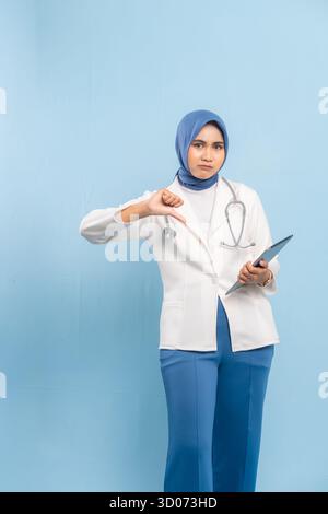Unzufriedene Ärztin in Hijab, die die Daumen unten Geste gibt, isoliert auf blauem Hintergrund Stockfoto