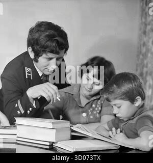 Eine sowjetische Familie versammelt sich um einen Tisch mit Büchern zu Hause in Sloviansk, 1970er bis 1980er Archivfoto Schwarzweiß, UdSSR, Ukrainische SSR. Der Vater, ein Maschinenführer in seiner Eisenbahnuniform, hilft seinem Sohn, ein Buch zu lesen. Die Mutter sitzt neben ihnen und beobachtet. Dieses herzerwärmende Porträt fängt eine Szene des sowjetischen Alltags, der Familienwerte, der Bildung zu Hause und der engagierten Erziehung ein. Die Uniform des Mannes unterstreicht die Verbindung der Familie zum Eisenbahnberuf. Ein nostalgisches Dokument des friedlichen Lebens im Donbass vor dem Krieg. Stockfoto