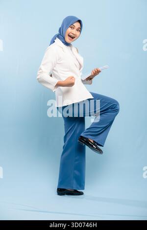 Porträt einer selbstbewussten asiatischen muslimischen Geschäftsfrau oder Studentin, die Stärke oder Entschlossenheit zeigt, lächelnd, isoliert auf blauem Hintergrund. Stockfoto