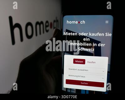 In dieser Fotoabbildung hält eine Person ein Smartphone mit der Webseite der Schweizer Immobilienportalgesellschaft Home.ch auf dem Bildschirm vor dem Logo. Stockfoto