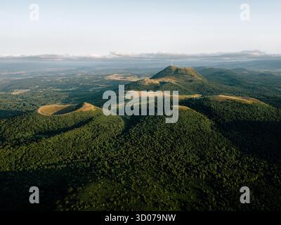 Luftbild des Chaîne des Puys in der Auvergne, Frankreich, mit vulkanischen Kratern, dem Gipfel des Puy de Dôme, dichten Wäldern und sanftem Sonnenuntergang über einem Tra Stockfoto