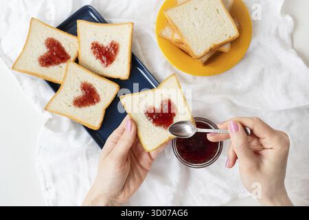 Das Mädchen macht einen Toast, auf dem das Herz aus Marmelade besteht. Überraschungsfrühstück im Bett. Romantik zum Valentinstag Stockfoto