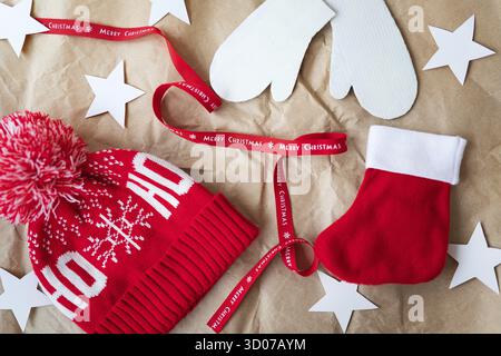 Rotes Band mit der Aufschrift Frohe Weihnachten zusammen mit einer roten Kappe, Fäustlinge, rote Socke auf Bastelpapier ausgebreitet. Weihnachtsfeier Konzept auf De Stockfoto