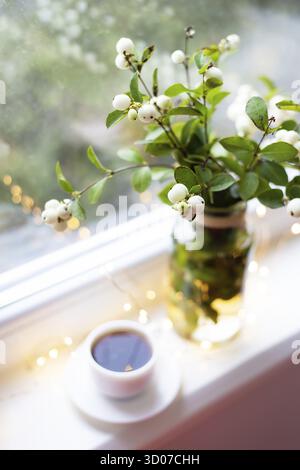 Eine weiße Schneebeere in einem Bouquet steht mit einer Tasse Kaffee auf der Fensterbank nahe dem Fenster, schöne Lichter, Bokeh im Fenster Stockfoto