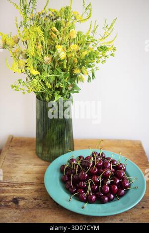 Ein lebhaftes Sommerstillleben mit einem Blumenstrauß aus gelben Wildblumen und einer Schüssel reifer roter Kirschen auf einem rustikalen Holztisch Stockfoto