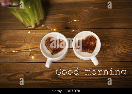 Zwei weiße Tassen Cappuccino stehen auf einem alten Holztablett. Garland, schön blinkend. Draufsicht, Nahaufnahme. Die Inschrift Guten Morgen Stockfoto