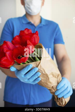Berührungslose Blumenlieferung per Kurier in Schutzmaske, medizinische Handschuhe mit einem Strauß roter Tulpen. Nahaufnahme Stockfoto
