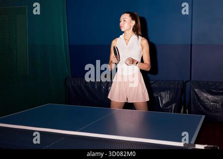 Kaukasische junge Erwachsene Frau, die sich darauf vorbereitet, während des Tischtennisspiels zu dienen, mit dem Paddel in der Hand in der Nähe des Tischtennistisches in der Sporthalle, fokussiert auf den Gegner zu stehen Stockfoto