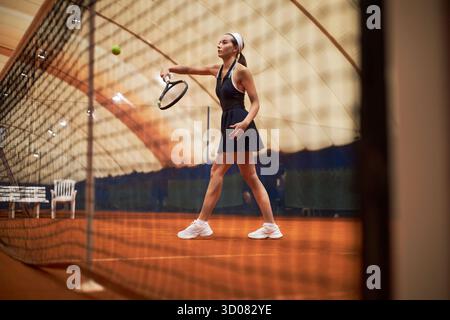 Junge Erwachsene Kaukasierin, die Tennis auf dem Hallenplatz spielt, Schläger hält und sich auf den Ball vorbereitet, athletisches Outfit trägt, sich auf das Spiel konzentriert, Netz im Vordergrund Stockfoto