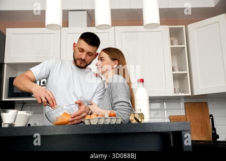 In einer modernen Küche teilen sich ein Mann und eine Frau einen Moment, während sie eine Mahlzeit zubereiten, Eier miteinander rühren und sich gegenseitig anlächeln, was ein warmes AT erzeugt Stockfoto