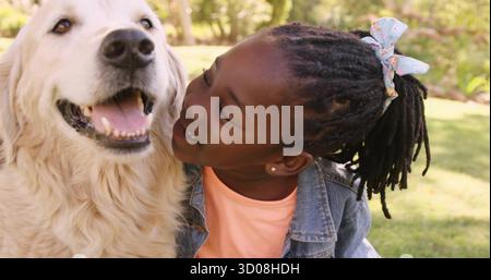 Umarmtes, geflochtenes Mädchen in Denim-Jacke, das sich im sonnendurchfluteten Park zum goldenen Retriever neigt, mit Blumenschleife Stockfoto
