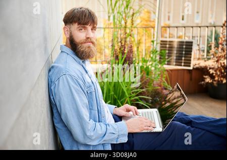 Porträt eines glücklichen bärtigen Mannes arbeitet auf dem Laptop auf dem Schoß. Solarpanel in der Nähe, der nachhaltigen, modernen, umweltfreundlichen Arbeitsbereich betont, der Technologie und Nachhaltigkeit vereint. Stockfoto