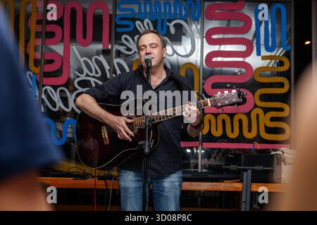 Ein Weißer mittleren Alters mit kurzen Haaren singt in ein Mikrofon, während er eine Akustikgitarre spielt. Farbenfroher Hintergrund mit abstrakten Designs. Stockfoto