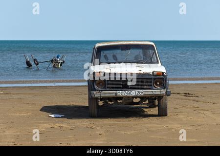 Buzowna, Aserbaidschan - 29. März 2025: Ein verwittertes weißes Vintage-Auto aus dem Jahr 1987 Lada Niva sitzt am Sandstrand, während blaue Wellen auf den Horizont treffen Stockfoto