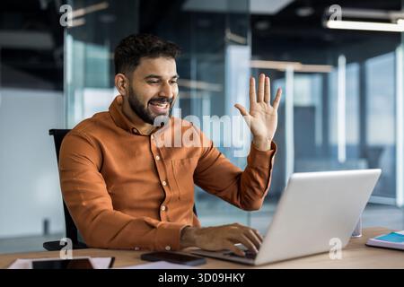 Indischer Geschäftsmann, der an einer Online-Konferenz mit einem Laptop am Schreibtisch teilnimmt, mit der Hand winkt, aktiv kommuniziert und Konzepte über virtuelle Kommunikation weitergibt Stockfoto