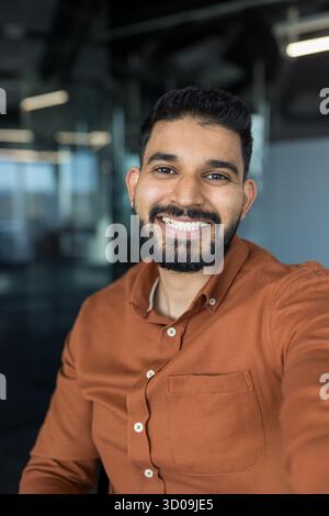 Lächelnder indischer Geschäftsmann mit Bart, der ein Selfie macht, einen glücklichen und positiven Ausdruck zeigt, selbstbewusst in einem modernen Büro oder Coworking Space steht und Erfolg und Professionalität repräsentiert Stockfoto