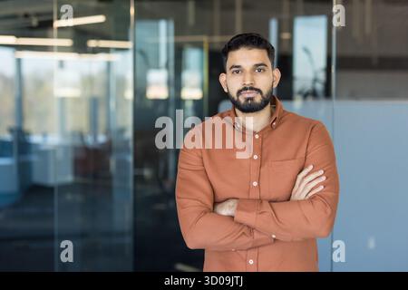 Selbstbewusster indischer Geschäftsmann oder Unternehmer, der mit überkreuzten Armen steht und einen fokussierten und entschlossenen Ausdruck in einer hellen, modernen Büroumgebung darstellt Stockfoto