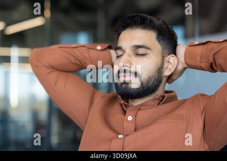 Junger bärtiger Mann in einem braunen Hemd, der sich in einem Büro entspannt, mit geschlossenen Augen und Händen hinter dem Kopf ausruht und während eines harten Arbeitstages Ruhe und Stressreduktion findet Stockfoto