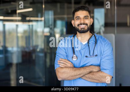 Junger indischer Gesundheitsexperte, der mit gekreuzten Armen steht, selbstbewusst lächelnd, mit blauen Peelings und einem Stethoskop in einer modernen medizinischen Umgebung Stockfoto
