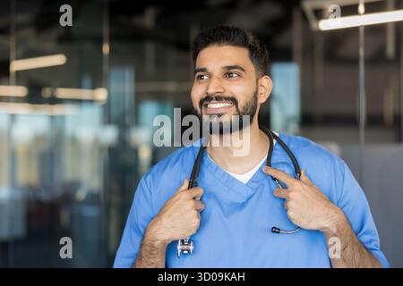 Junger indischer männlicher Mediziner, der blaue Peelings und ein Stethoskop um den Hals trägt, wegblickt und selbstbewusst lächelt in einem modernen Krankenhaus oder einer Klinik Stockfoto