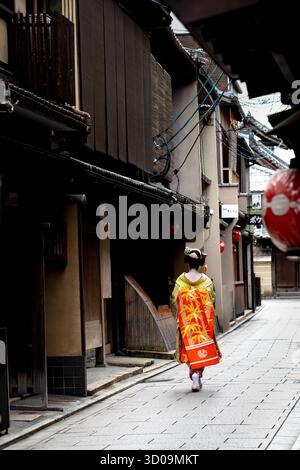 Ein traditionell gekleidetes Maiko geht in Kyoto, Japan, eine ruhige Straße entlang, umgeben von hölzernen Machiya-Stadthäusern und roten Laternen. Stockfoto