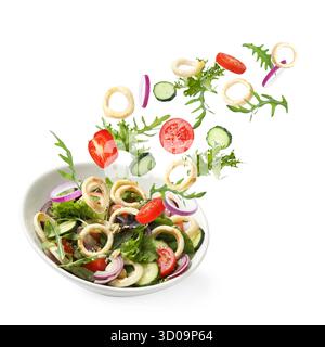 Tintenfischringe und andere Zutaten, die in eine Schüssel mit Salat vor weißem Hintergrund fallen Stockfoto