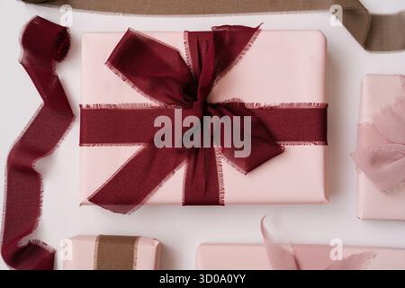 Flache Lagen eleganter rosafarbener Geschenkboxen, umwickelt mit weichen Stoffbändern in Burgunderrot- und staubigen Rosatönen auf weißem Hintergrund. Minimalistische Ästhetik Stockfoto